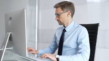 Modern bir ofisteki bir bilgisayarda çalışan işadamı, masasında yazı yazarken görevlerine odaklanıyor, profesyonelliği, verimliliği ve kurumsal bir ortamda kendini adamayı somutlaştırıyor.