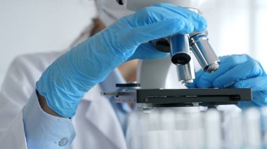 Mikroskobu ayarlayan eldivenli bilim adamlarına yakın çekim yapmak, laboratuvarda araştırma yapmak ve arka planda mavi sıvı içeren test tüpleri kullanmak. Tıp, sağlık ve bilim konsepti.