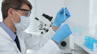Laboratuvar önlüklü, koruyucu gözlüklü ve mavi eldivenli maskeli erkek bilim adamı modern laboratuardaki test tüplerine mikroskop ve test tüpleri rafı koyuyor. Tıp, sağlık ve bilim konsepti.