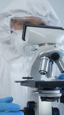 Tehlikeli madde giysisi, maske ve mavi eldiven giyen erkek bir virüs bilimci laboratuvarda virüsleri araştırırken mikroskop altında özenle çalışıyor. Tıp ve bilim konsepti.