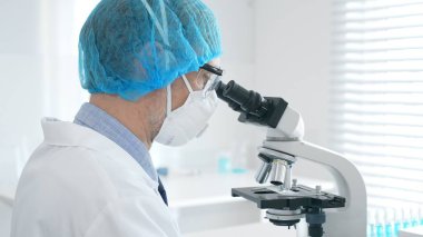 Üst düzey erkek bilim adamı koruyucu maske, saç filesi, gözlük ve eldiven takıyor mikroskop kullanarak tıbbi bir laboratuarda bilimsel araştırma yapıyor. Laboratuvar ve bilim konsepti
