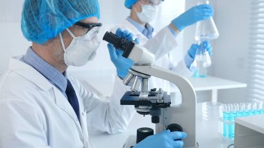 Koruyucu maske takan ve gözlük takan bilim adamı mikroskop kullanarak laboratuvarda araştırma yapıyor ve arka planda elinde mataralar olan bir bilim adamı asistanı var. Sağlık ve bilim konsepti.