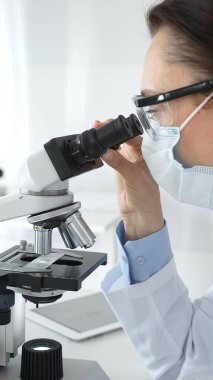 Koruyucu maskeli ve gözlüklü bir kadın bilim adamı modern bir laboratuvarda araştırma yaparken bir örneği mikroskop altında dikkatlice inceliyor. Tıp ve bilim kavramı.