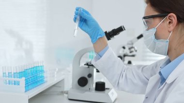 Koruyucu maske takan dişi araştırmacı ve mavi eldiven test tüpünde sıvıyı inceliyor, laboratuvarda mikroskop ve laboratuvar ekipmanlarının yanında oturuyor. Tıp, sağlık ve bilim konsepti.
