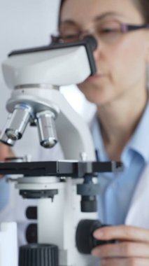 Koruyucu ekipman takan kadın bilim araştırmacısı mikroskobu kontrol ediyor, modern laboratuvar araştırma ortamında mavi test tüplerini analiz ediyor. Tıp ve bilim konsepti.