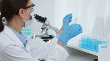 Yüz maskesi takan ve mavi eldiven takan kadın bir bilim adamı modern bir laboratuarda bir deney tüpüne dikkatlice sıvı bırakıyor. Tıp ve bilim konsepti.