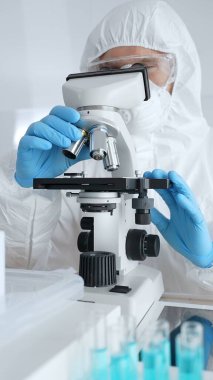 Koruyucu ekipman takan bilim araştırmacısı mikroskobu ayarlıyor, laboratuvar örneklerini steril araştırma ortamında hassasiyetle analiz ediyor. Tıp, sağlık ve bilim konsepti.