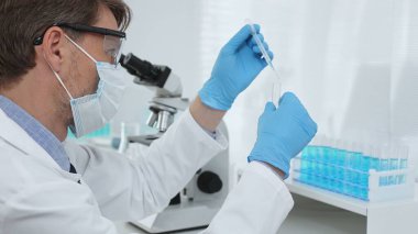 Laboratuvar önlüklü, koruyucu gözlüklü ve mavi eldivenli maskeli erkek bilim adamı modern laboratuardaki test tüplerine mikroskop ve test tüpleri rafı koyuyor. Tıp, sağlık ve bilim konsepti.