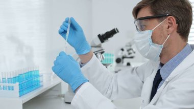 Laboratuvar önlüklü, koruyucu gözlüklü ve mavi eldivenli maskeli erkek bilim adamı modern laboratuardaki test tüplerine mikroskop ve test tüpleri rafı koyuyor. Tıp, sağlık ve bilim konsepti.