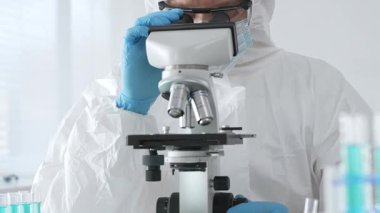 Tehlikeli madde giysisi giyen tıp virolojisti laboratuvarda mikroskopla çalışıyor. Yeni virüsler için potansiyel tedavileri araştırırken mavi sıvı örneklerini inceliyor.