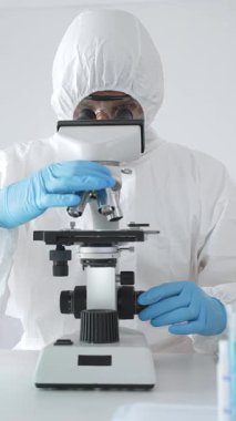 Tehlikeli madde giysisi giyen tıp virolojisti laboratuvarda mikroskopla çalışıyor. Yeni virüsler için potansiyel tedavileri araştırırken mavi sıvı örneklerini inceliyor.