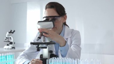Beyaz laboratuvar önlüğü giyen bilim araştırmacısı mikroskop ile numune inceliyor, test tüpleriyle steril laboratuvar ortamında biyolojik araştırmalar yapıyor.