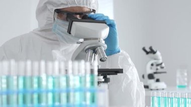 Laboratuvar ortamında araştırma yaparken mikroskop kullanarak koruyucu kıyafet giyen bir tıp bilimcisi. Test tüplerini mavi sıvıyla kapatın..