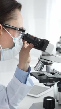 Koruyucu maske takan ve gözlük takan kadın bilim adamı mikroskopla numuneleri dikkatle inceliyor ve modern laboratuvarda araştırma yapıyor. Tıp ve bilim konsepti.