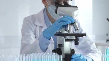 Mikroskobu ayarlayan, laboratuardaki örnekleri inceleyen, yeni ilaç veya kimyasal formülasyon araştırmaları yapan laboratuvar önlüğü giyen ve güvenlik için yüz maskesi takan bilim insanlarının eldiveni kapalı.