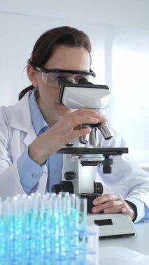 Beyaz laboratuvar önlüğü giyen bilim araştırmacısı mikroskop ile numune inceliyor, test tüpleriyle steril laboratuvar ortamında biyolojik araştırmalar yapıyor.