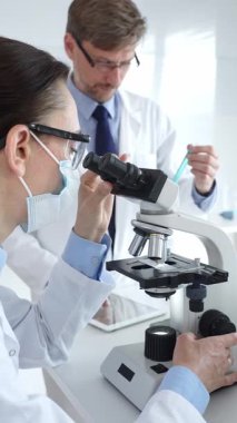 Koruyucu gözlük ve maske takan kadın bilim adamı, laboratuvarda tıbbi araştırma sırasında mikroskoptan bakıyor ve erkek meslektaşı arka planda çalışıyor. Tıp ve Bilim