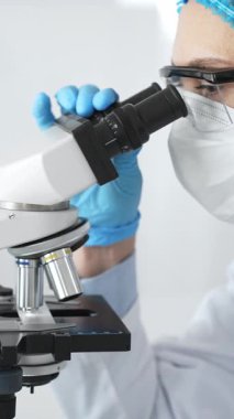 Mavi koruyucu giysiler giyen kadın bilim adamı mikroskop kullanarak laboratuvar ortamında örnekler inceleyerek bilimsel araştırma ve analiz yapıyor. Tıp ve bilim konsepti.