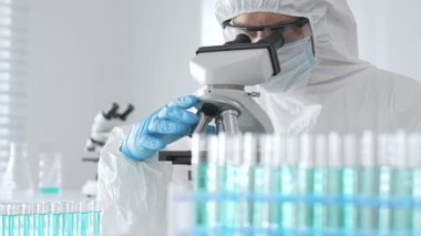 Laboratuvar ortamında araştırma yaparken mikroskop kullanarak koruyucu kıyafet giyen bir tıp bilimcisi. Test tüplerini mavi sıvıyla kapatın..
