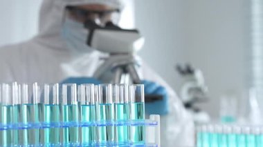 Laboratuvar ortamında araştırma yaparken mikroskop kullanarak koruyucu kıyafet giyen bir tıp bilimcisi. Test tüplerini mavi sıvıyla kapatın..