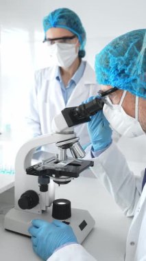 Laboratuvar önlüklü, yüz maskeli ve koruyucu gözlüklü iki bilim adamı laboratuarda mikroskop kullanarak araştırma yapıyorlar. Tıp ve bilim konsepti.