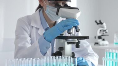 Mikroskobu ayarlayan, laboratuardaki örnekleri inceleyen, yeni ilaç veya kimyasal formülasyon araştırmaları yapan laboratuvar önlüğü giyen ve güvenlik için yüz maskesi takan bilim insanlarının eldiveni kapalı.