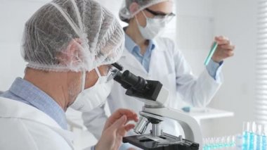 Koruyucu maske takan kıdemli erkek bilim adamı, gözlük ve saç filesi takıyor, mikroskop kullanarak genç bayan meslektaşlarına yardım ediyor, tablet tutuyor ve laboratuvarda notlar alıyor. Tıp ve bilim konsepti.