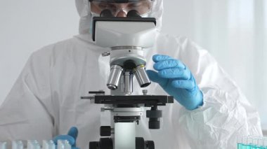 Tehlikeli madde giysisi giyen tıp virolojisti laboratuvarda mikroskopla çalışıyor. Yeni virüsler için potansiyel tedavileri araştırırken mavi sıvı örneklerini inceliyor.