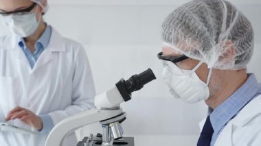 Koruyucu maske takan kıdemli erkek bilim adamı, gözlük ve saç filesi takıyor, mikroskop kullanarak genç bayan meslektaşlarına yardım ediyor, tablet tutuyor ve laboratuvarda notlar alıyor. Tıp ve bilim konsepti.