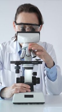 Beyaz laboratuvar önlüğü giyen bilim araştırmacısı mikroskop ile numune inceliyor, test tüpleriyle steril laboratuvar ortamında biyolojik araştırmalar yapıyor.