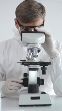 Laboratuvar önlüğü ve gözlüklü bir tıp bilimcisi örnekleri incelerken mikroskop kullanarak laboratuvarda araştırma yapıyor..