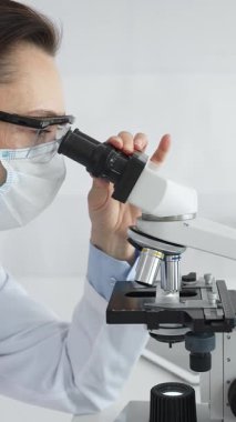 Koruyucu maske takan ve gözlük takan kadın bilim adamı mikroskopla numuneleri dikkatle inceliyor ve modern laboratuvarda araştırma yapıyor. Tıp ve bilim konsepti.