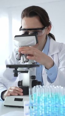 Beyaz laboratuvar önlüğü giyen bilim araştırmacısı mikroskop ile numune inceliyor, test tüpleriyle steril laboratuvar ortamında biyolojik araştırmalar yapıyor.