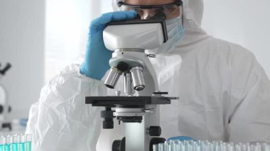 Tehlikeli madde giysisi giyen tıp virolojisti laboratuvarda mikroskopla çalışıyor. Yeni virüsler için potansiyel tedavileri araştırırken mavi sıvı örneklerini inceliyor.