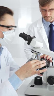 Koruyucu gözlük ve maske takan kadın bilim adamı, laboratuvarda tıbbi araştırma sırasında mikroskoptan bakıyor ve erkek meslektaşı arka planda çalışıyor. Tıp ve Bilim.