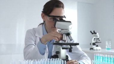 Beyaz laboratuvar önlüğü giyen bilim araştırmacısı mikroskop ile numune inceliyor, test tüpleriyle steril laboratuvar ortamında biyolojik araştırmalar yapıyor.