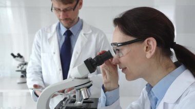 Laboratuvarda araştırma sırasında mikroskoptan bakan koruyucu gözlük takan kadın bir bilim adamı ve arka planda çalışan erkek arkadaşı. Tıp ve bilim konsepti.