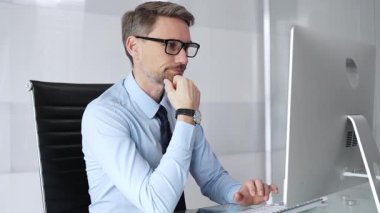 Temiz bir ofis masasında oturan, gözlüklü, kravatlı, açık mavi gömlekli bir iş adamı, bilgisayar üzerinde düşünüyor ve çalışıyor..