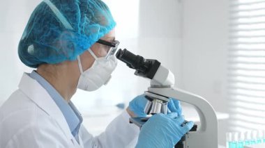 Kadın araştırmacı mikroskop ile numune inceliyor, steril laboratuvar içinde çalışma yerini çevreleyen cam şişelerle tıbbi deney yapıyor. Tıp ve Bilim.