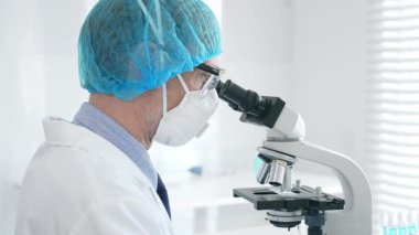 Üst düzey erkek bilim adamı koruyucu maske, saç filesi, gözlük ve eldiven takıyor mikroskop kullanarak tıbbi bir laboratuarda bilimsel araştırma yapıyor. Laboratuvar ve bilim konsepti