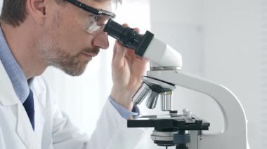 Yetişkin erkek bilim araştırmacısı mikroskopla örnek analiz ediyor, yüksek teknoloji laboratuvar ortamında koruyucu giysiler giyiyor. Sağlık, tıp ve bilim konsepti.