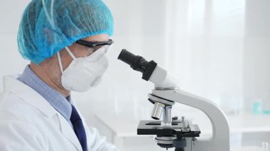 Üst düzey erkek bilim adamı koruyucu maske, saç filesi, gözlük ve eldiven takıyor mikroskop kullanarak tıbbi bir laboratuarda bilimsel araştırma yapıyor. Laboratuvar ve bilim konsepti