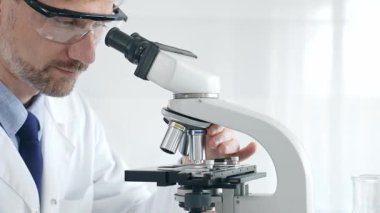 Yetişkin erkek bilim araştırmacısı mikroskopla örnek analiz ediyor, yüksek teknoloji laboratuvar ortamında koruyucu giysiler giyiyor. Sağlık, tıp ve bilim konsepti.