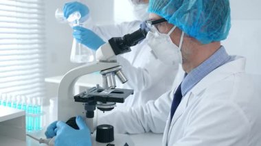 Koruyucu maske takan ve gözlük takan erkek bilim adamı mikroskop kullanarak laboratuvarda araştırma yapıyor ve elinde mataralar olan bir bilim adamı asistanı var. Sağlık ve bilim konsepti.
