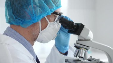 Koruyucu ekipman takan erkek bilim adamı mikroskop kaydırağını inceliyor, steril laboratuvar ortamında araştırma örneklerini analiz ediyor. Sağlık ve bilim konsepti.