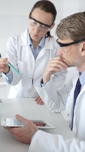 A male scientist takes notes on a tablet while his female colleague helps him looks through a microscope in a laboratory. Concept of medicine and science. A male scientist takes notes on a tablet while his female colleague helps him looks through a microscope in a laboratory. Concept of medicine and science.