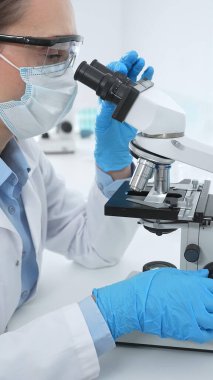 Mavi koruyucu giysili kadın bilim adamı laboratuvar araştırma ortamında test örneklerini mikroskop altında analiz ediyor. Tıp, sağlık ve bilim konsepti.