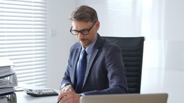 Takım elbiseli, kravatlı ve gözlüklü olgun girişimci masada oturuyor, notlar yazıyor ve modern, parlak ofiste hesap makinesi kullanıyor. Ticarette denetim ve vergiler.