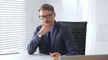 Mavi takım elbiseli ve kuyruklu yetişkin işadamı, parlak ve modern bir ofiste çalışırken konsantre oluyor ve düşünüyor. Profesyonelliğini ve verimliliğini gösteriyor. İş adamları konsepti.