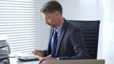 Takım elbiseli ve kravatlı olgun bir iş adamı ofis masasında oturuyor ve hesap makinesi kullanarak evrakları imzalıyor. İş dünyasında denetim ve vergi teması.